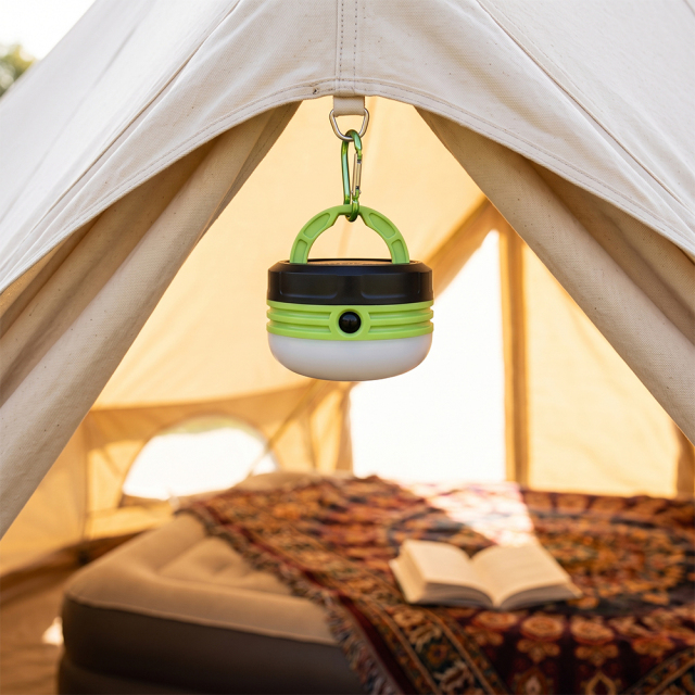 A green mini LED camping lantern hanging inside a outdoor tent above a mattress and book.