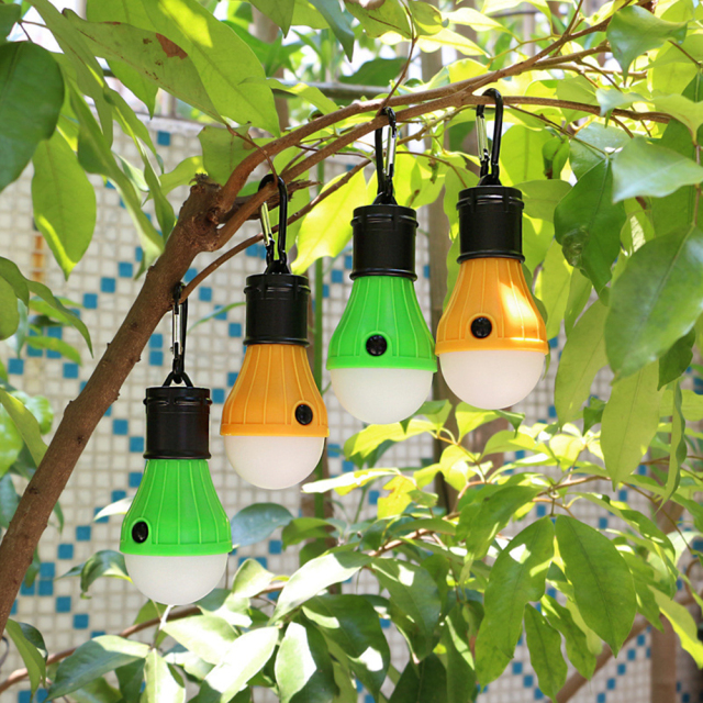 Four colorful LED camping bulb lights hanging from green tree branches outdoors.