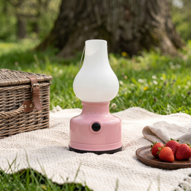 A pink vintage LED camping lantern sitting on a picnic blanket in the grass next to strawberries and a basket.