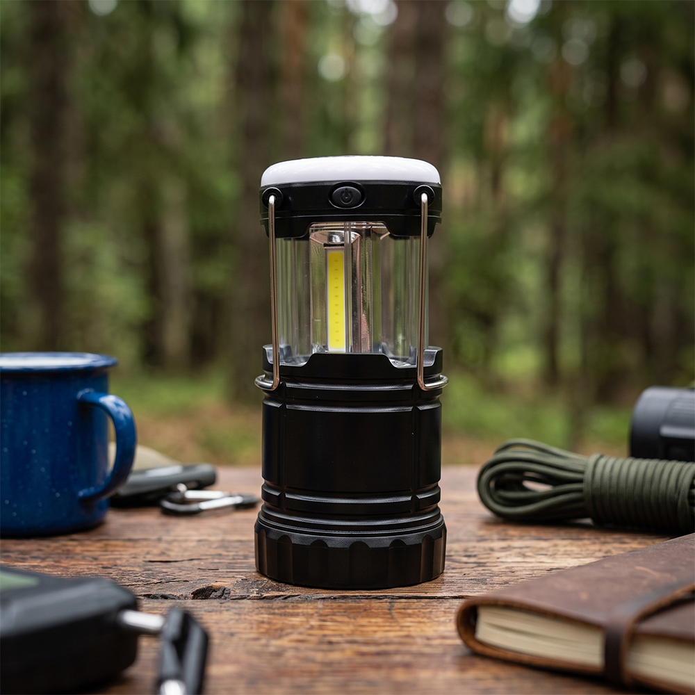 A glowing collapsible COB LED camping lantern on a wooden picnic table with an outdoor blurred background.