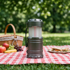 A dark grey collapsible LED flame camping lantern placed on a red and white checkered picnic mat with fruits and a basket.