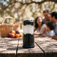 A black collapsible LED flame camping lantern sitting on a wooden picnic table with a family in the blurred background.