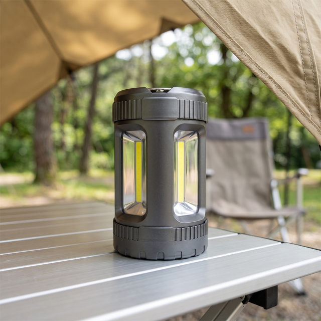A black rugged COB LED camping lantern placed on an aluminum folding camping table near a tent.