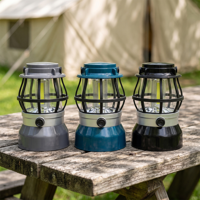Three vintage COB LED camping lanterns in grey, blue, and black placed on a wooden outdoor table near a tent.