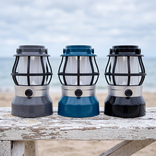 Three vintage LED flame camping lanterns in grey, blue, and black displayed on a white wooden table at the beach.