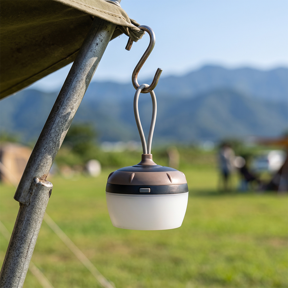 A mini portable LED tent light hanging from an S-hook on a camping tent pole outdoors.