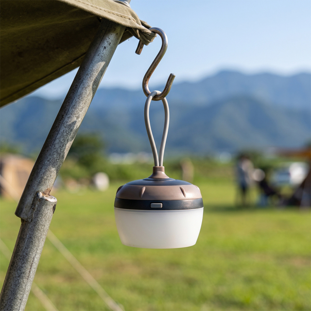 A mini portable LED tent light hanging from an S-hook on a camping tent pole outdoors.