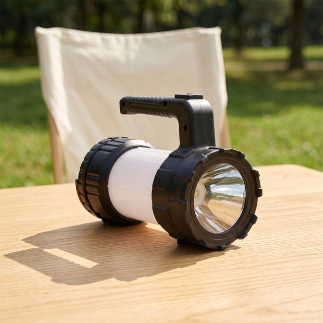 A black LED camping lantern searchlight sitting on a wooden table outdoors near a folding chair.