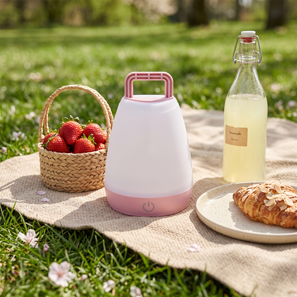 A pink portable LED camping light sitting on a picnic blanket on the grass next to strawberries and a drink bottle.
