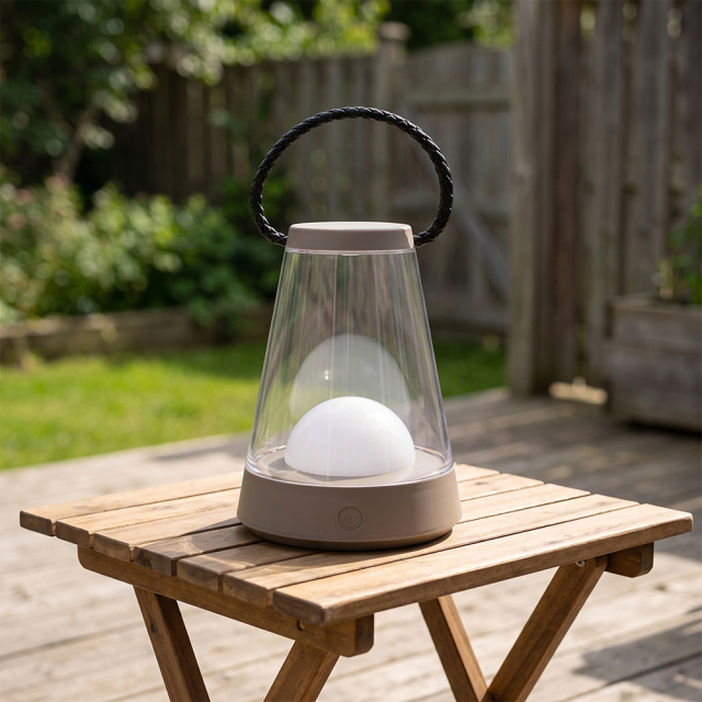 A portable LED camping lantern with a brown transparent shade and woven handle sitting on a wooden patio table.