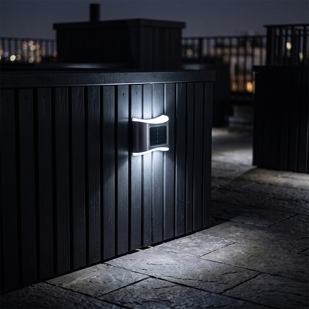 A stainless steel solar up and down fence light glowing with cool white light on a black wooden deck wall at night.