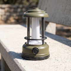 An army green vintage LED camping lantern sitting on an outdoor stone bench on a sunny day.