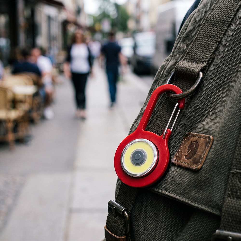 A red COB LED keychain light with carabiner clip attached to a canvas backpack outdoors.