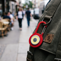 A red COB LED keychain light with carabiner clip attached to a canvas backpack outdoors.