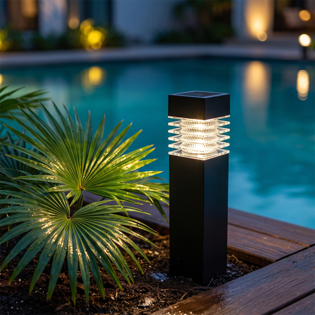 A black solar bollard light illuminating a pool deck surrounded by green palm leaves at night.