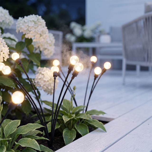 Solar firefly stake lights glowing warmly in a flowerbed beside a white wooden patio deck.