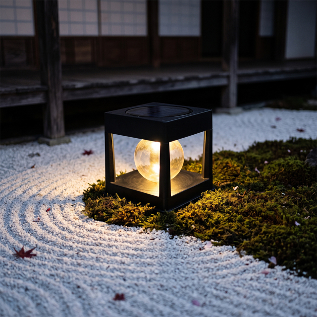 A black solar pathway lantern light glowing warmly on a mossy border beside raked white zen garden gravel.