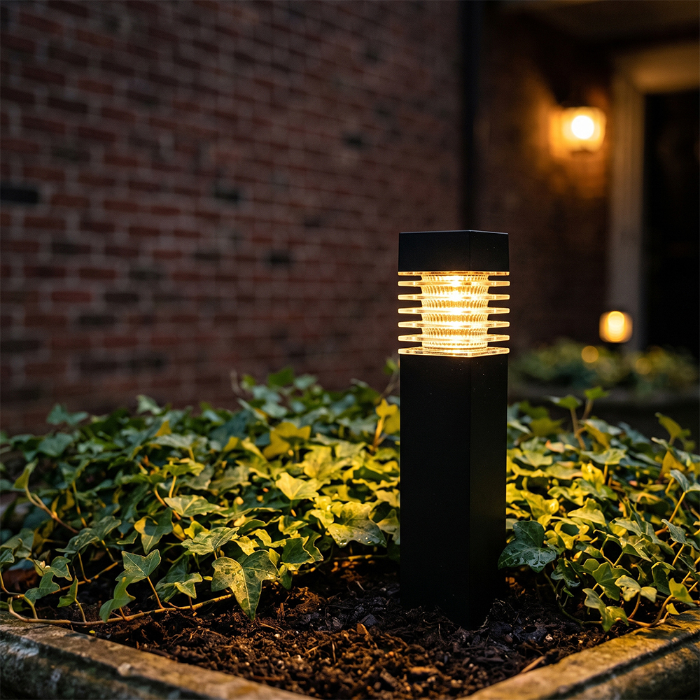 A solar pathway bollard light shining brightly in a garden bed with green ivy near a red brick wall.