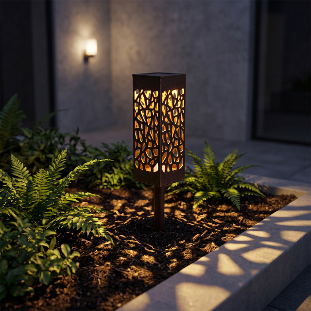 A black hollowed-out solar pathway light glowing in a modern garden flowerbed at night.