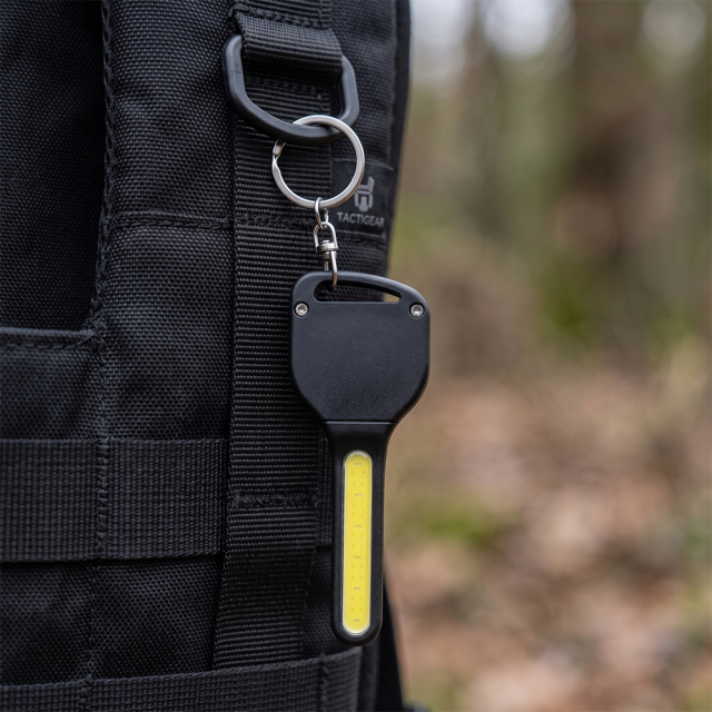 A key-shaped COB LED keychain light attached to a black outdoor backpack ring.