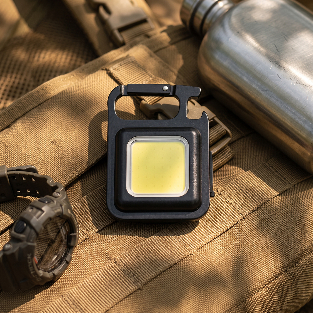 A black rechargeable COB keychain light placed on a tactical backpack next to a water bottle.
