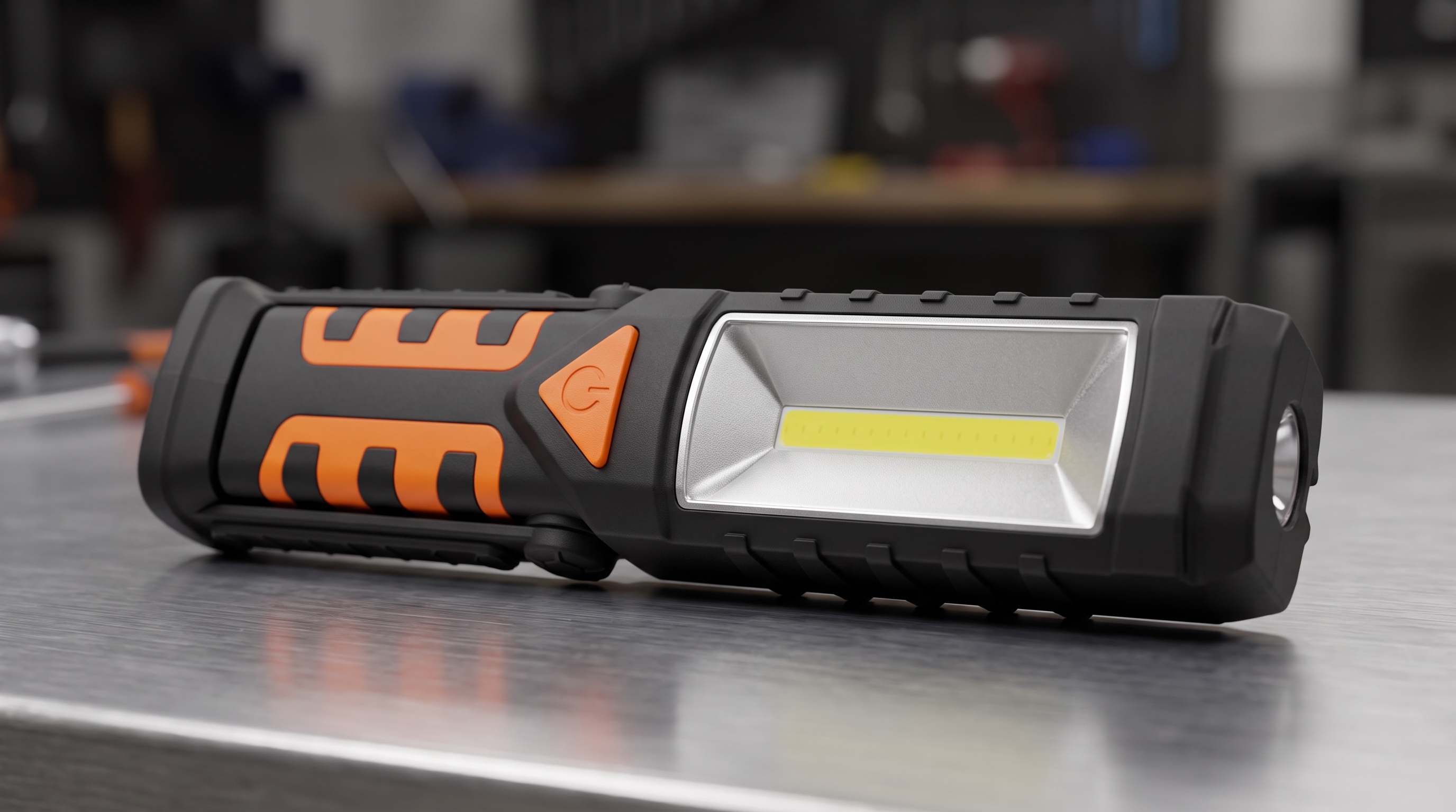 Black and orange portable COB LED work light with side spotlight lying horizontally on a metal workbench in a professional workshop.