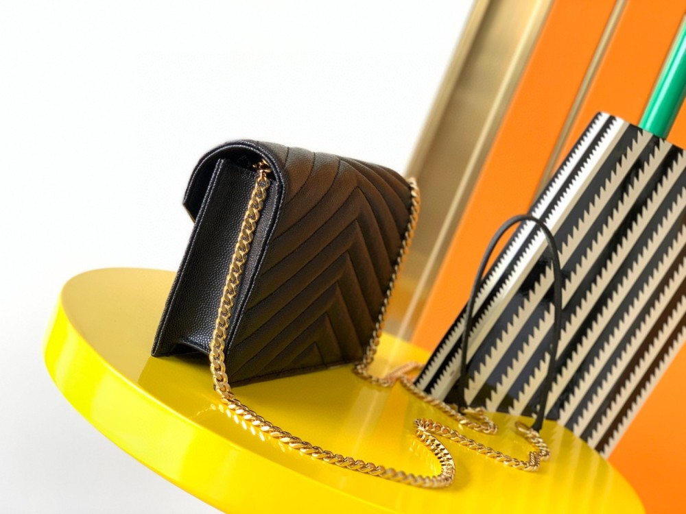 This is the YSL Cassandre chain wallet, crafted from black grained leather with quilted detailing and a gold-tone metal YSL logo on the front. It can be worn as a shoulder or crossbody bag, combining practicality with brand recognition.