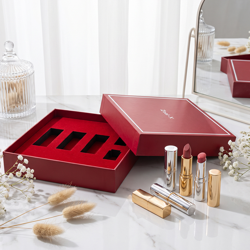 Red lid-and-base makeup gift box with multiple insert compartments displayed beside gold cosmetic tubes