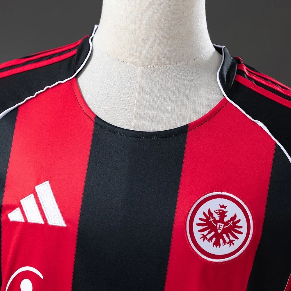Close-up of the Eintracht Frankfurt 2025-26 home jersey showing the embroidered Adidas logo and club crest on the red and black striped fabric.