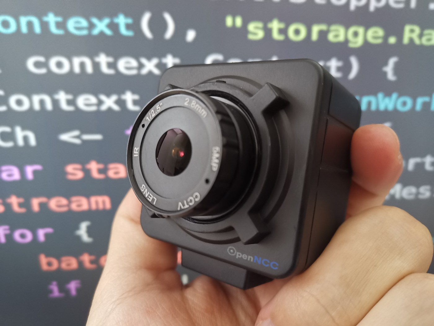 Hand holding the OpenNCC Knight USB AI camera in front of a screen with source code - the OpenNCC-branded enclosed camera runs Intel OpenVINO models on-board